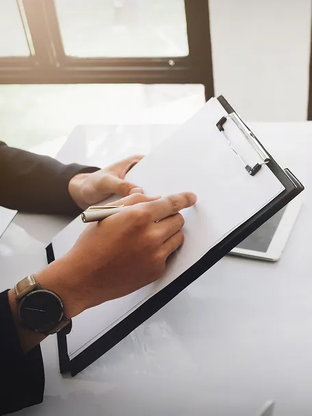 Eine Hand hält einen Stift und schreibt auf ein Klemmbrett, während ein Tablet im Hintergrund sichtbar ist.