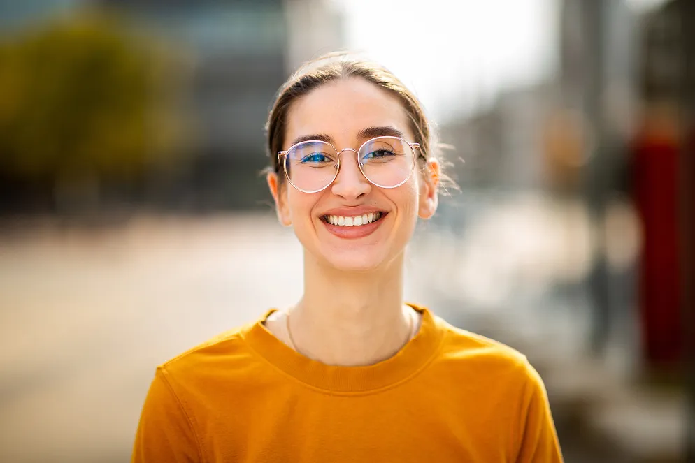 Lächelnde Person mit Brille in orangefarbenem Shirt, unscharfer urbaner Hintergrund.
